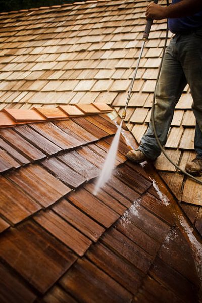 Man stands atop a roof while spraying the wooden shingles clean. Vertical shot.